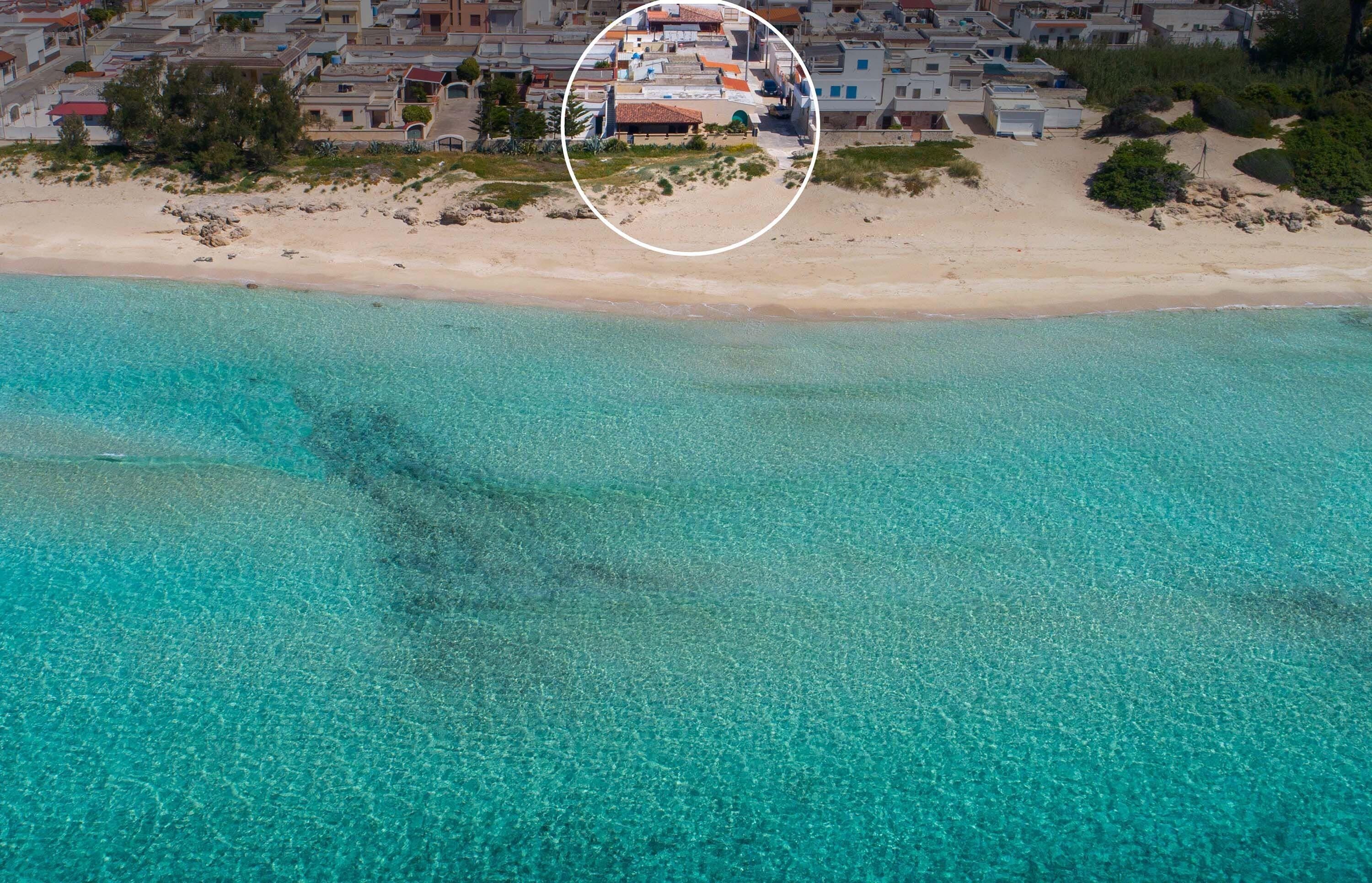An aerial view of a coastal area with clear turquoise waters, a sandy beach, and a cluster of buildings with a distinct structure highlighted by a circle.