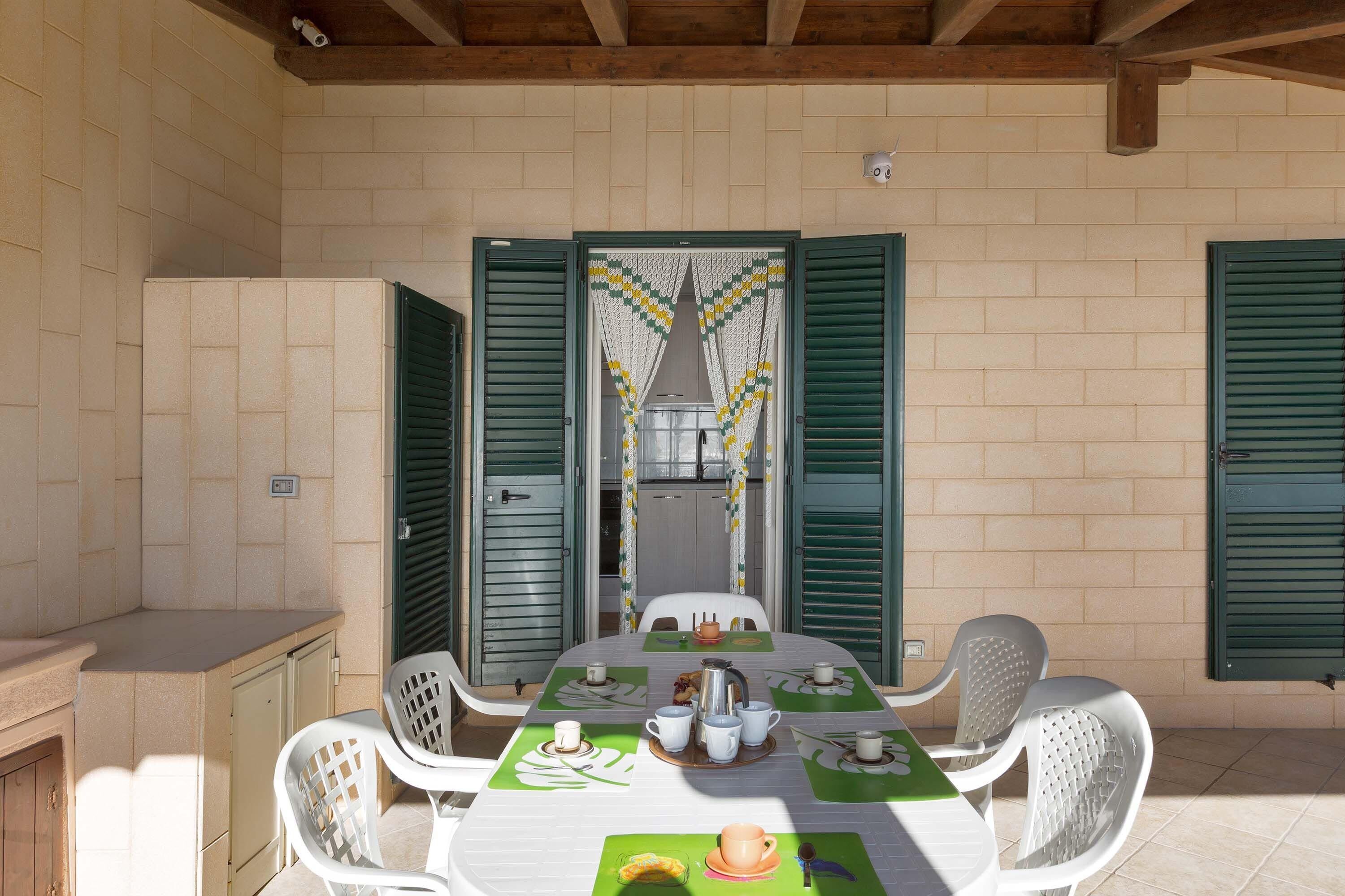 Un espacio de comedor al aire libre con techo, una mesa dispuesta para cuatro personas, sillas de plástico blanco alrededor, y contraventanas verdes en las paredes.