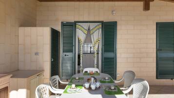 Un espacio de comedor al aire libre con techo, una mesa dispuesta para cuatro personas, sillas de plástico blanco alrededor, y contraventanas verdes en las paredes.