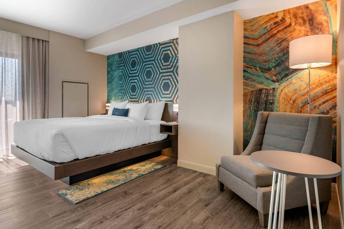 A modern hotel room with a large bed, a seating area with a small table, and a decorative wall with geometric patterns.