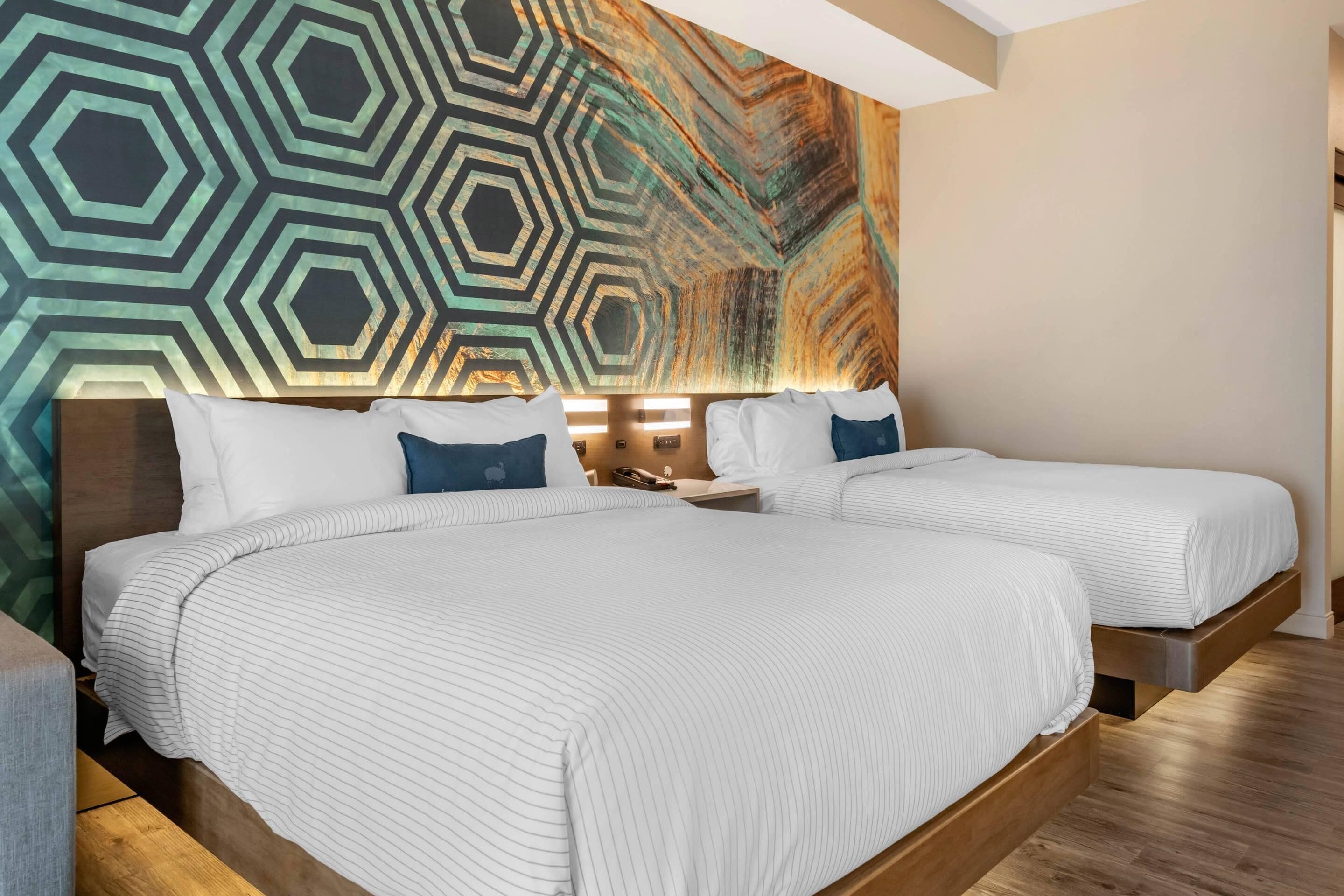 A hotel room with two beds, a geometric wall mural, and a wooden floor.
