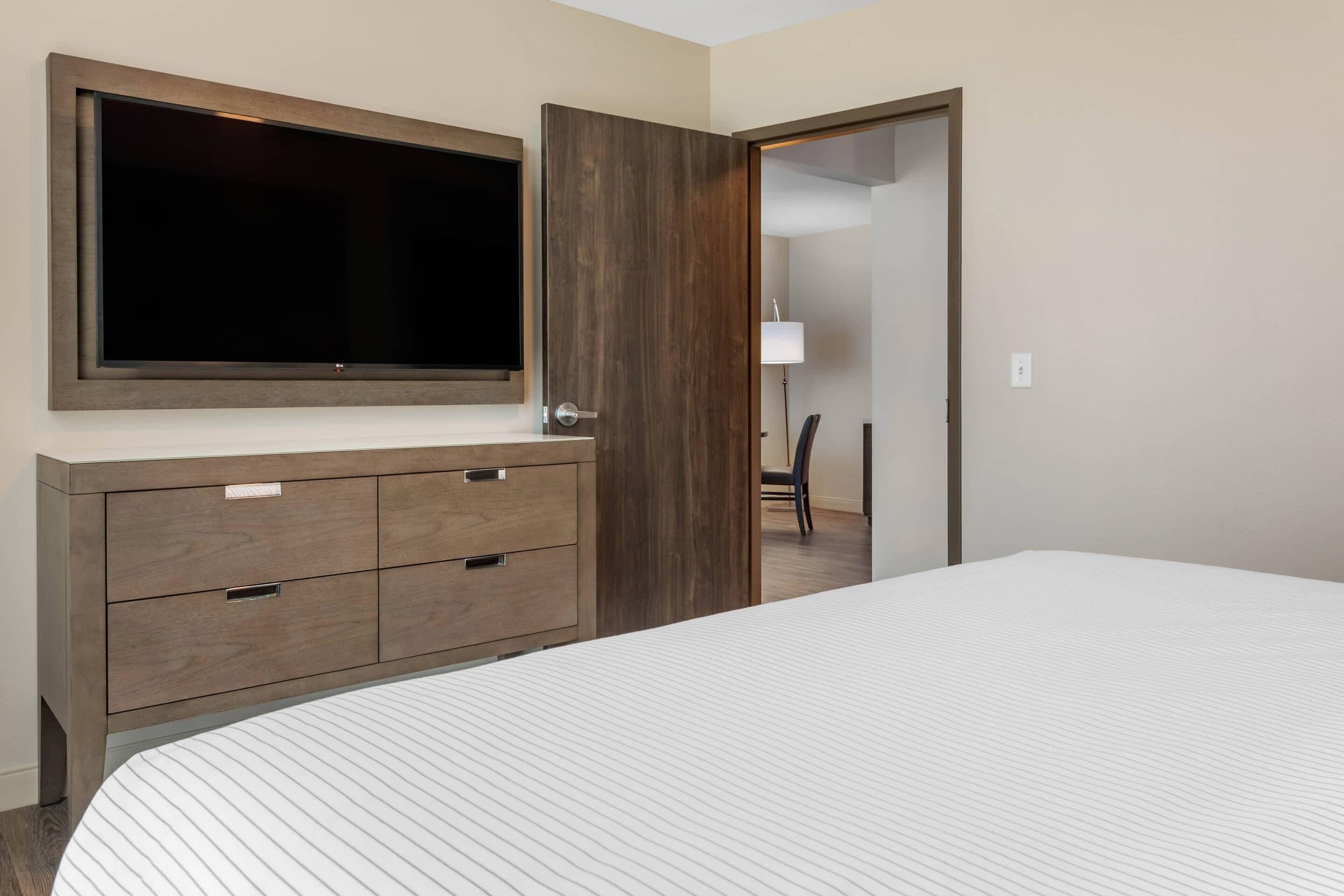 Una habitación de hotel con una cama grande, un aparador de madera con cajones, un televisor de pantalla plana montado sobre el aparador, y una puerta que da a otra habitación.