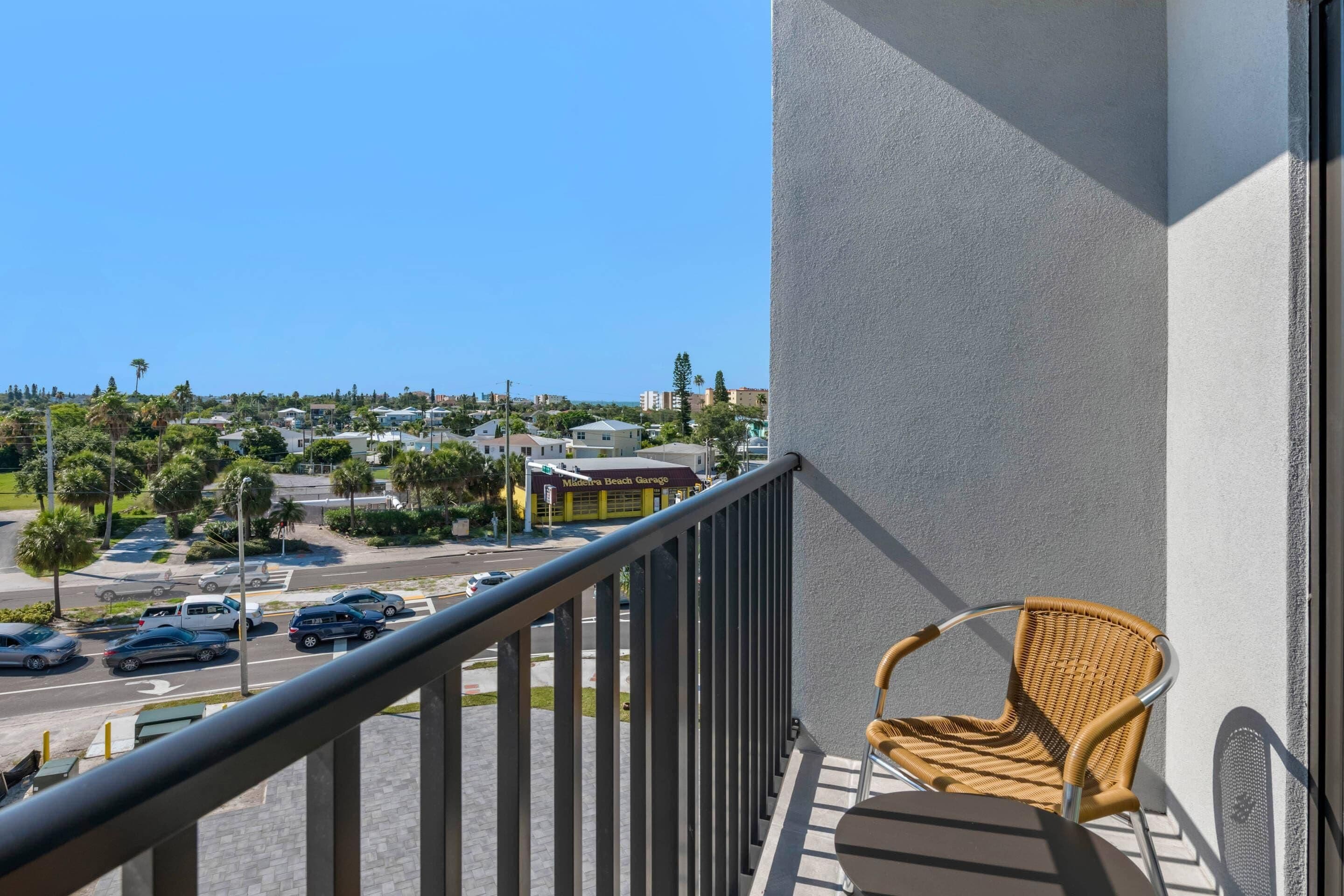 A balcony with a wicker chair, a clear view of a street with cars and buildings, and a clear blue sky.