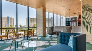 A modern lounge area with a blue sofa, wire chairs, and a round table. Large windows offer a view of palm trees and buildings. - Cambria Hotel St Petersburg-Madeira Beach Marina (Madeira Beach)
