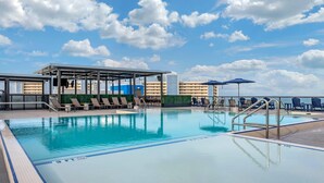 A swimming pool with lounge chairs and umbrellas, surrounded by a covered area with a view of the city skyline. - Cambria Hotel St Petersburg-Madeira Beach Marina (Madeira Beach)