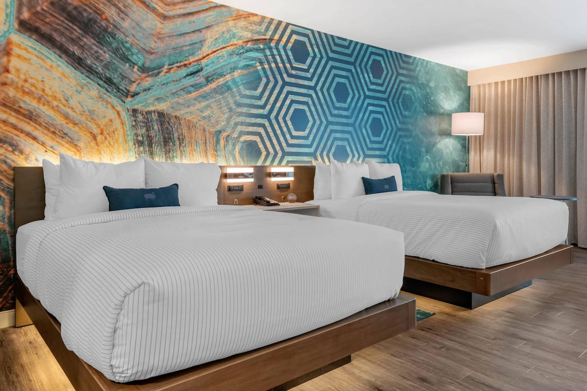 A hotel room with two beds, a large abstract mural on the wall, a wooden floor, and a modern lamp.