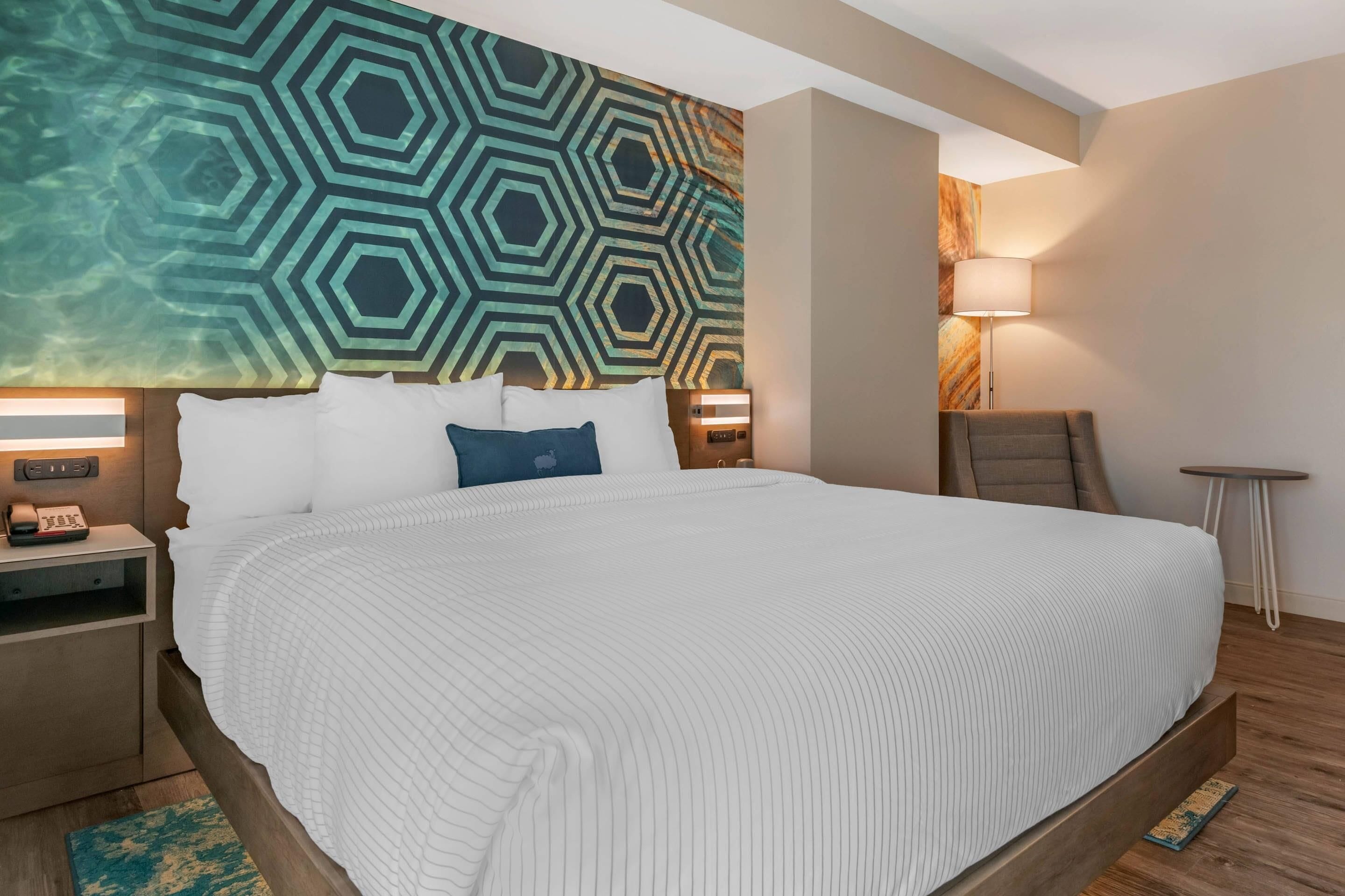 Une chambre d’hôtel moderne avec un grand lit, un mur orné d’un motif géométrique, une table de chevet avec une lampe et une petite table d’appoint.