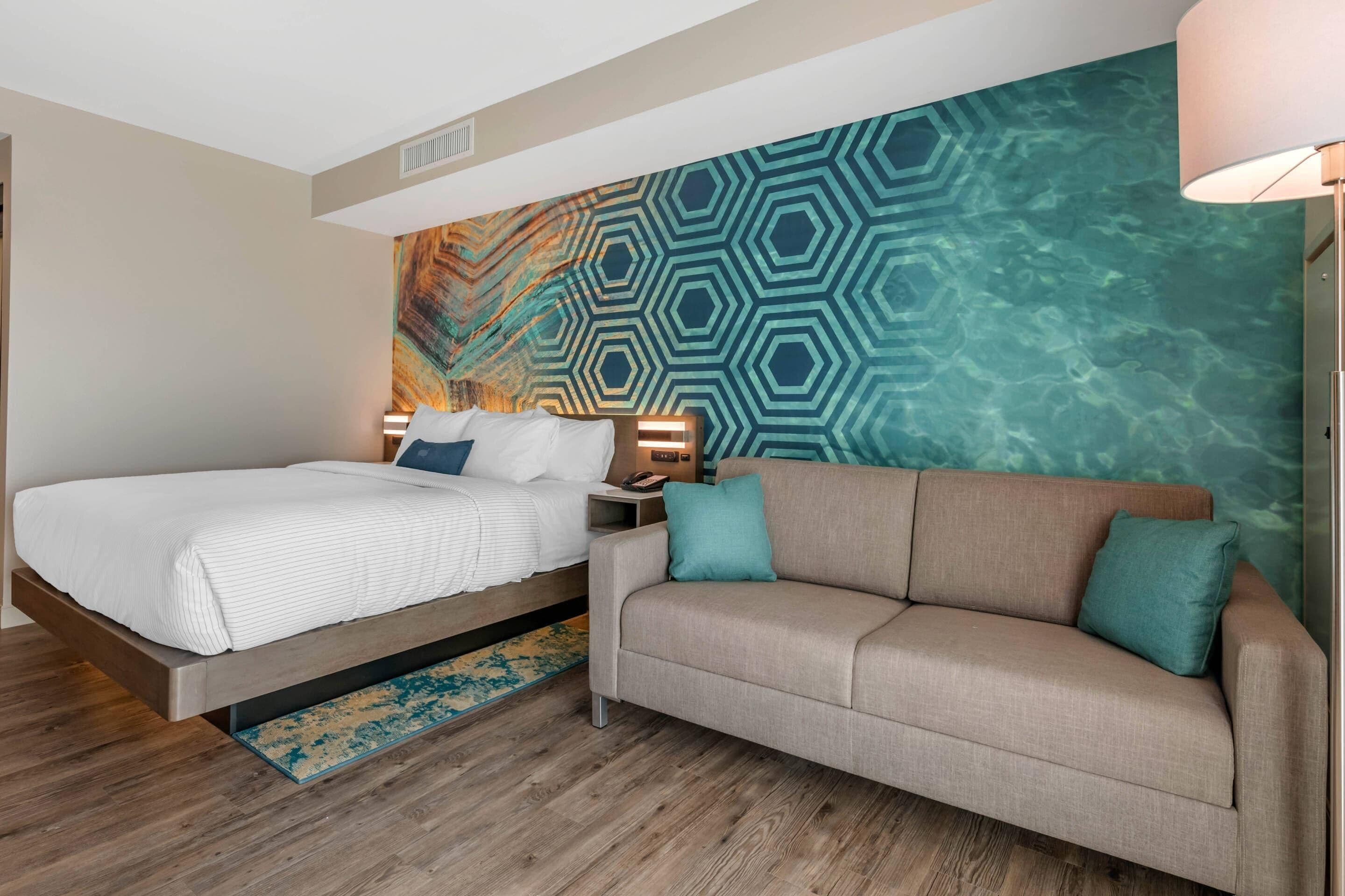 Una habitación de hotel con cama, sofá, mesita de noche y pared con diseño geométrico.