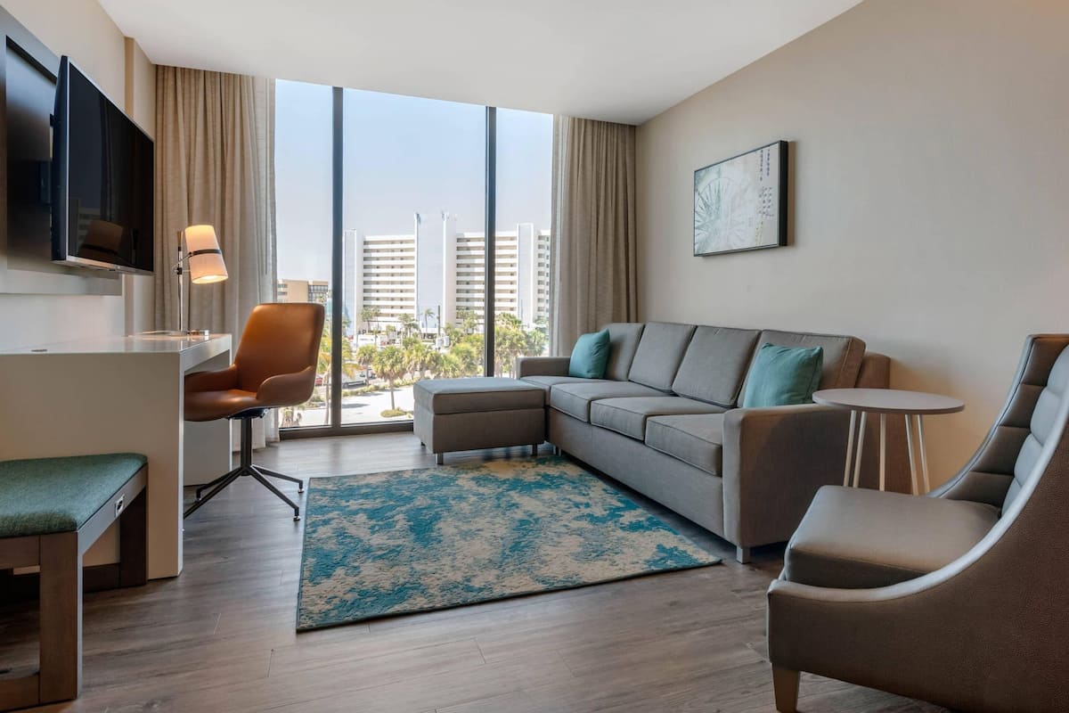 A modern hotel room with a large window, a sofa, a chair, a small table, a desk, and a flat-screen TV.