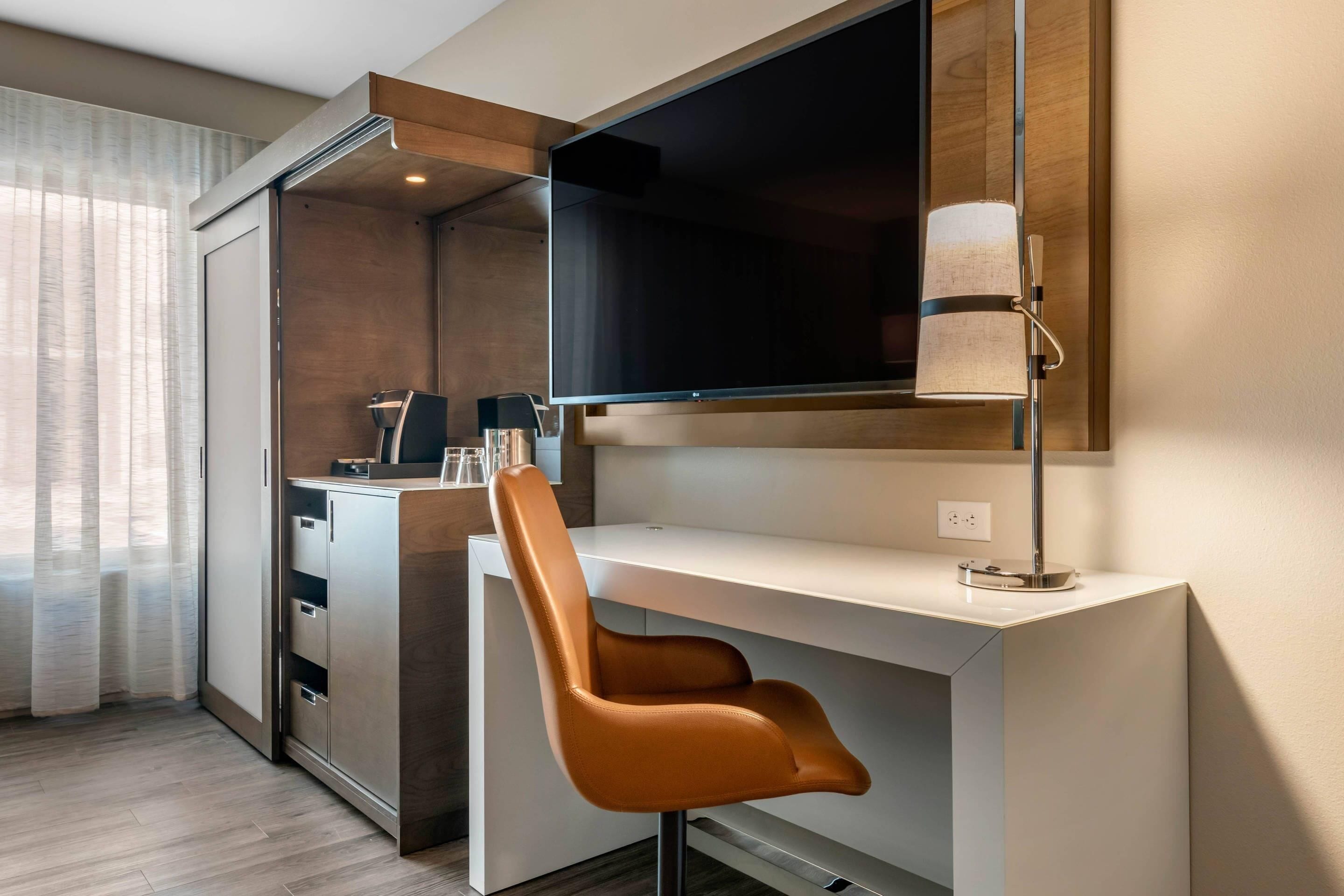 Ein modernes Hotelzimmer mit weißem Schreibtisch, braunem Ledersessel, Flachbildfernseher an der Wand und einer Lampe.