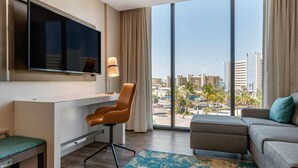 A hotel room with a large TV, a desk, a brown chair, a grey sofa, and a view of the city. - Cambria Hotel St Petersburg-Madeira Beach Marina (Madeira Beach)
