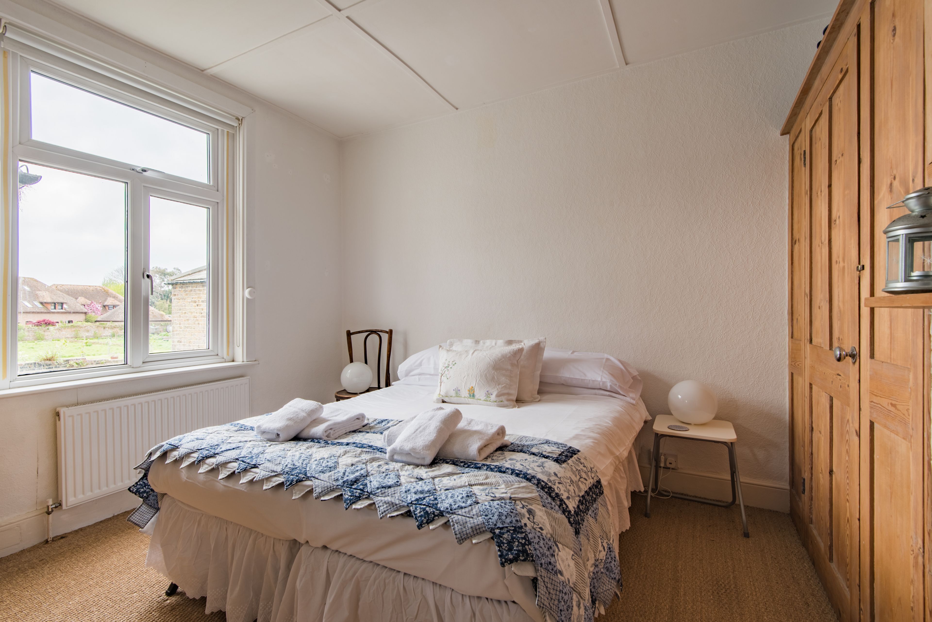 Een slaapkamer met een bed, een raam, een radiator, een houten kledingkast en een nachtkastje met een lamp.
