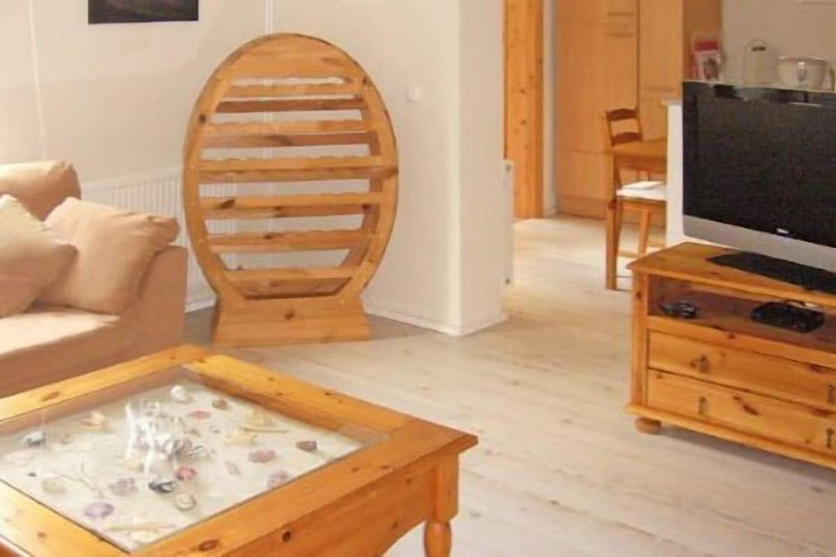 Una sala de estar con un sofá, una mesa de centro de madera con cubierta de vidrio, un televisor sobre un mueble de madera y una silla de madera.