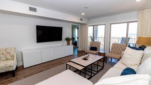 Uma sala de estar moderna com televisão de tela plana, um sofá, duas poltronas, uma mesa de centro e vista para a praia através de grandes janelas.