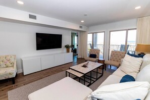 A modern living room with a flat-screen TV, a sofa, two armchairs, a coffee table, and a view of the beach through large windows. - SeeRay Shores 102 (St. Augustine)