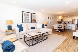 A modern living room with a sofa, coffee table, and a dining area with chairs and a kitchen in the background.