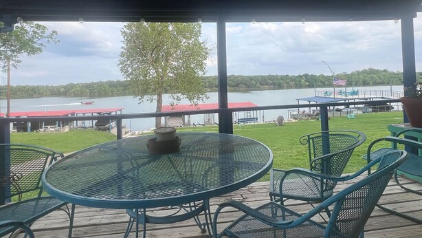 Un espace extérieur abrité, doté d’une table ronde et de chaises, qui offre une vue sur un lac où l’on peut apercevoir des bateaux et un quai.