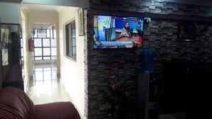 A room with a television displaying a news program, a sofa, and a wall-mounted TV. - Royal Palm Hotel And Suites (Nakuru)