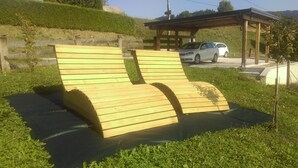 A wooden bench with a curved backrest and armrests, situated on a paved area with grass around it.