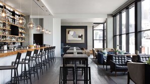 A modern bar area with a long counter, multiple seating options including bar stools and tables, and a large window offering a view of the city. - Canopy by Hilton Austin Downtown (Austin)