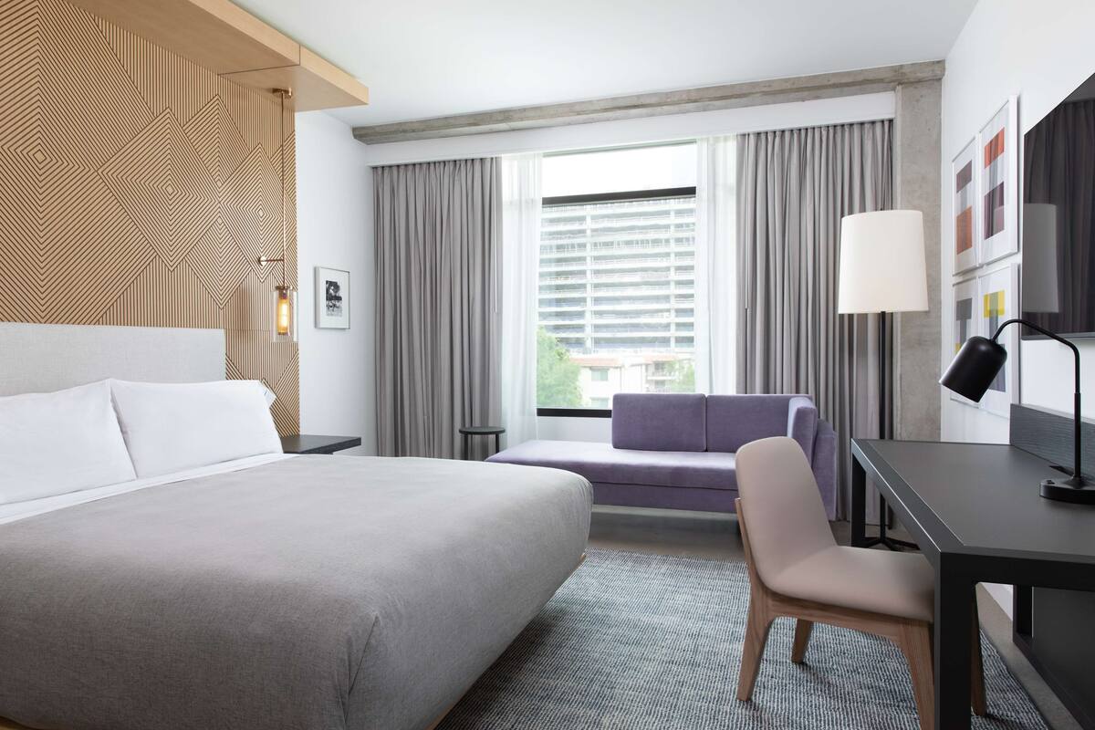 A hotel room with a large bed, a desk, a chair, a sofa, and a window with curtains.