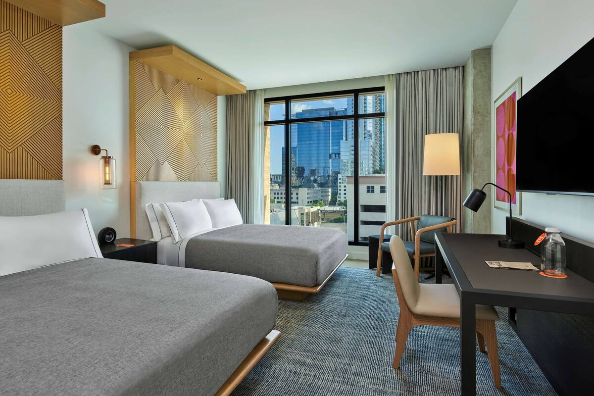 A hotel room with two beds, a desk, a chair, a TV, and a large window with a city view.