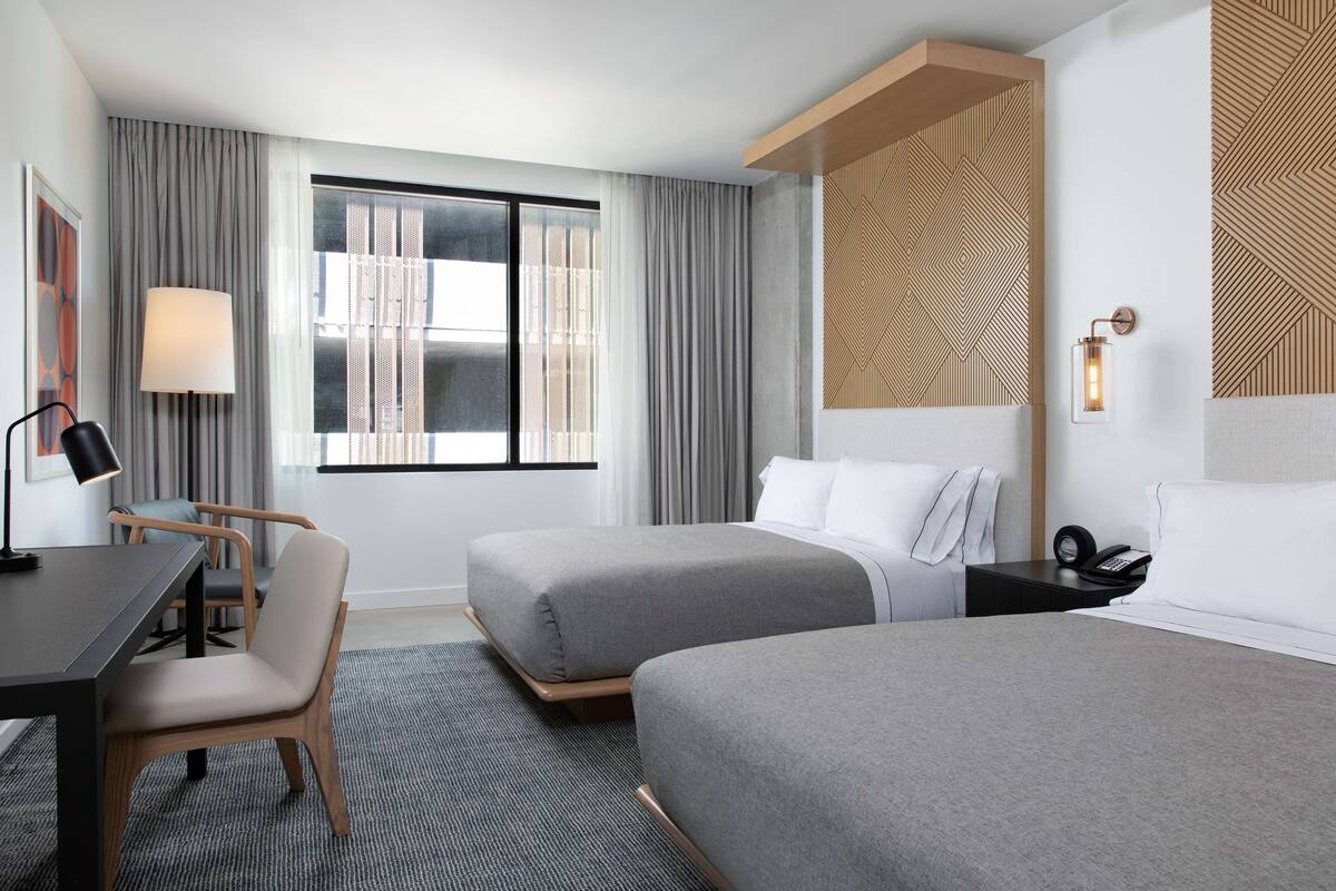 A hotel room with two beds, a desk, a chair, a lamp, and a window with curtains.