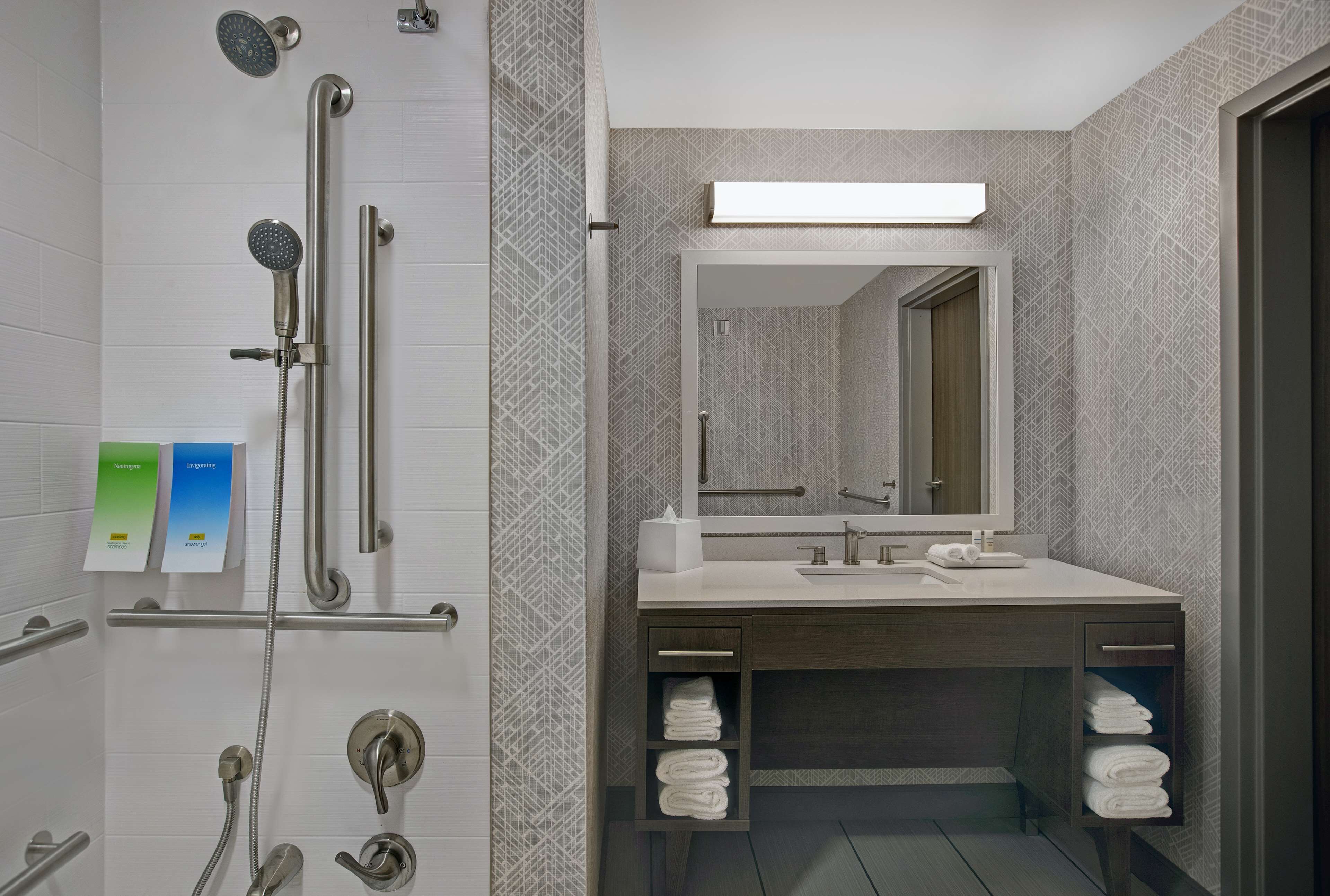 Une salle de bain moderne dotée d’une douche à l’italienne, de barres d’appui, d’un meuble-lavabo avec miroir et de porte-serviettes.