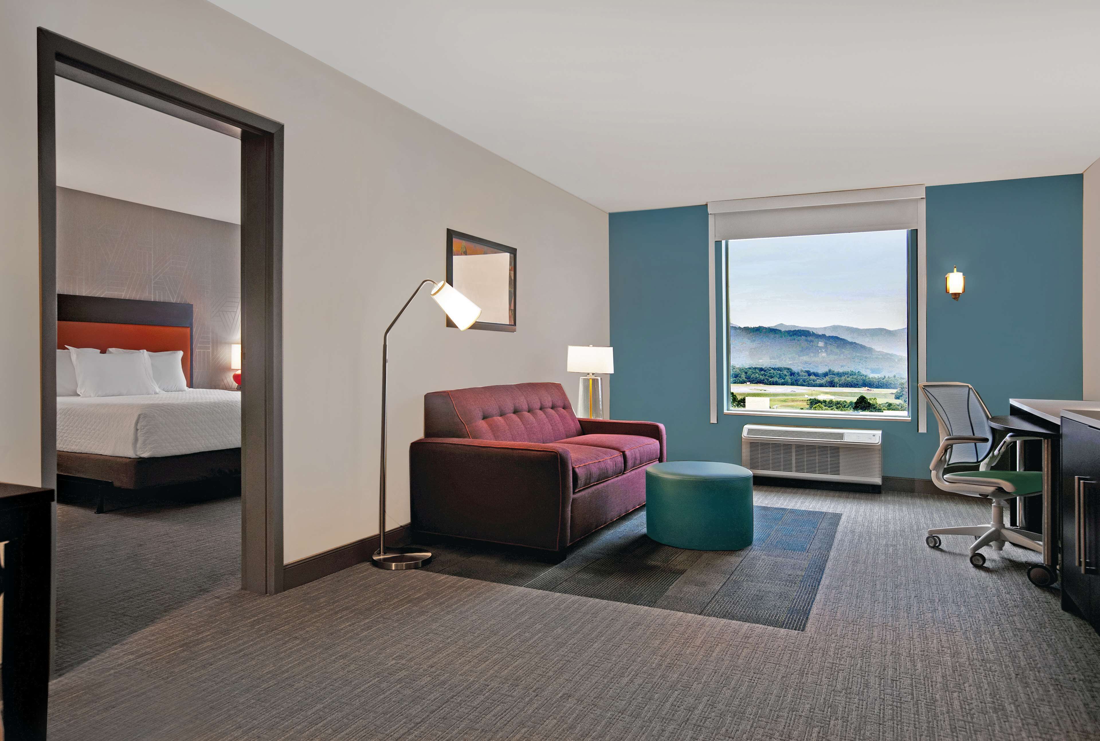 A hotel room with a bed, a red sofa, a desk, a chair, a standing lamp, and a large window with a mountain view.