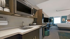 A modern hotel room with a kitchenette, a bed, a desk, and a view of the city.