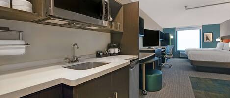 A modern hotel room with a kitchenette, a bed, a desk, and a view of the city.