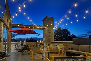 An outdoor patio area with string lights, a red umbrella, wicker furniture, and a stone pillar. - Home2 Suites Asheville Airport (Arden)