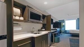 A modern kitchen with a microwave, sink, and cabinets. There is a small dining area with a chair and a table. The room has a large window and a bed in the background.