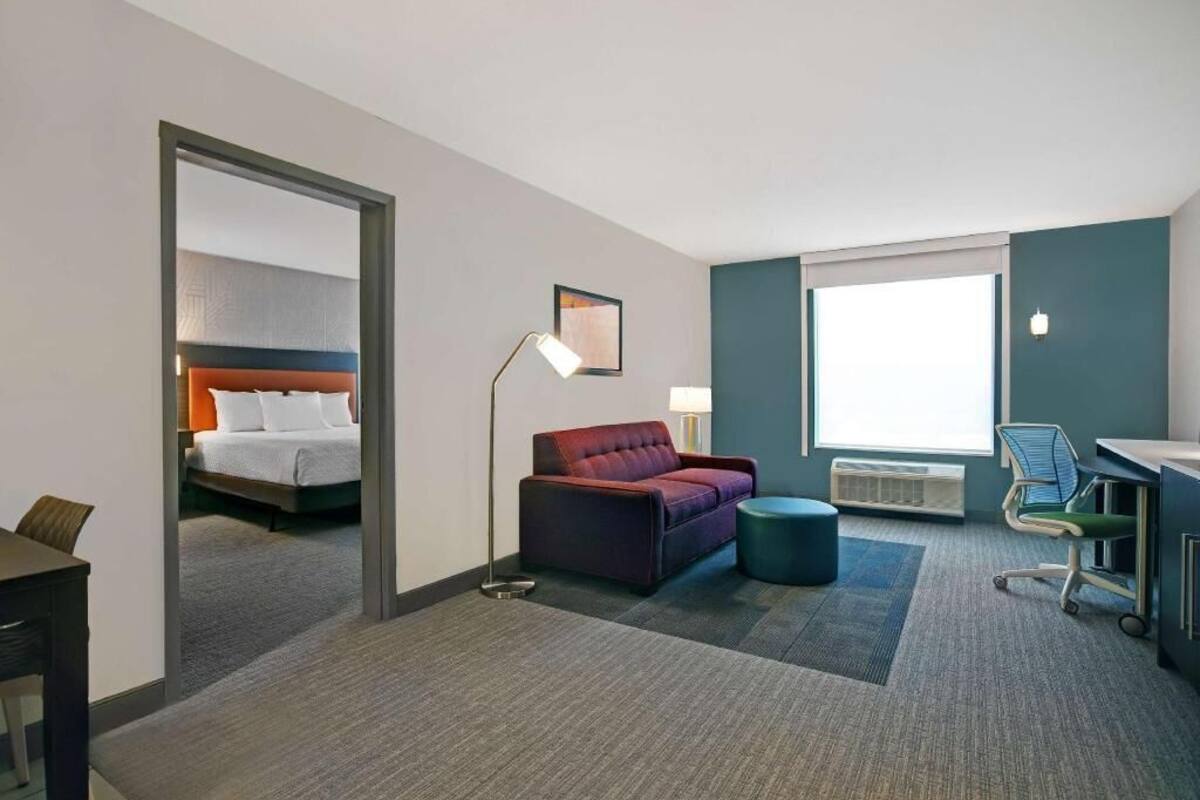 Ein Hotelzimmer mit einem Bett, einem Schreibtisch, einem Sessel, einer Couch, einer Lampe und einem großen Fenster.