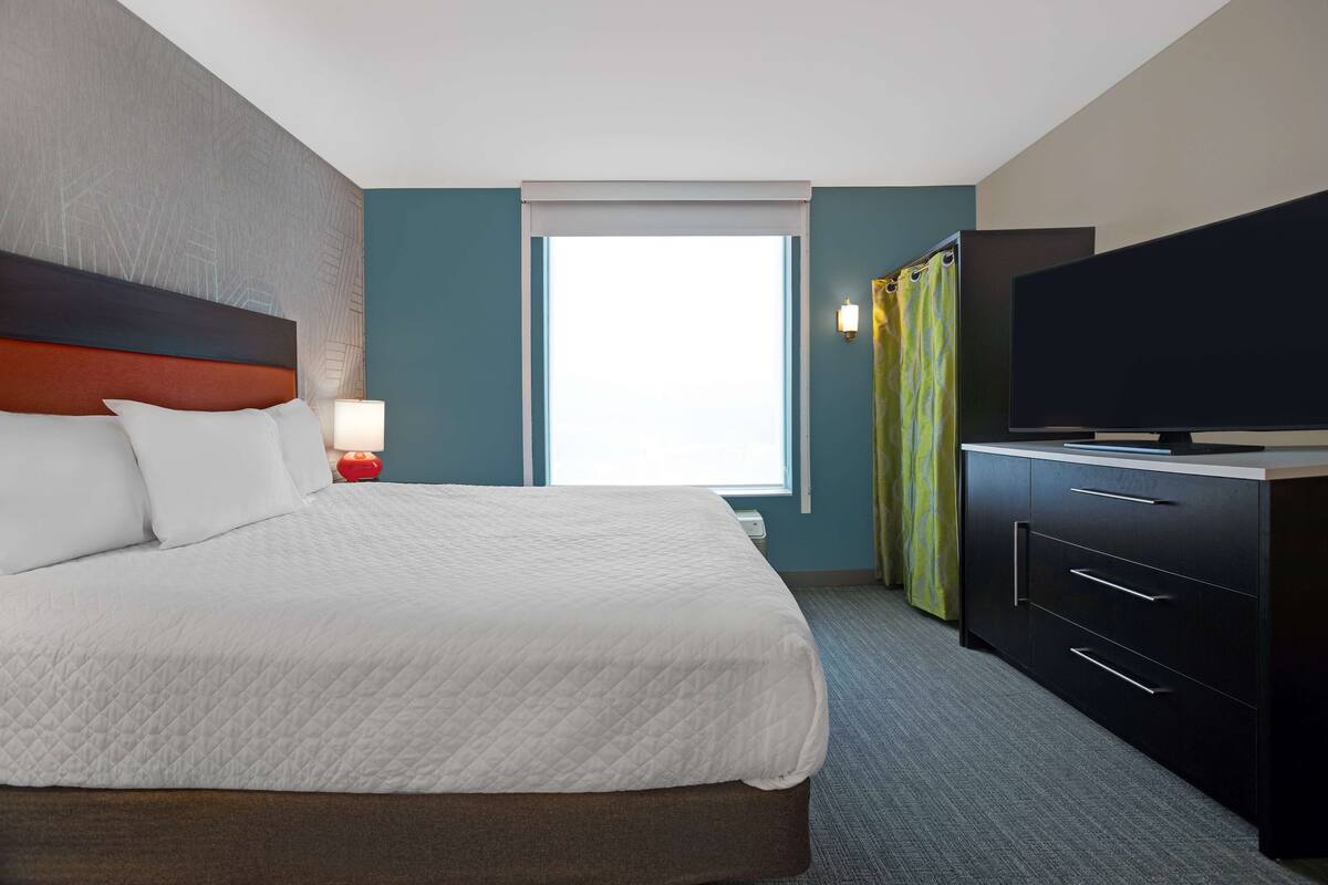 Ein Hotelzimmer mit einem großen Bett, einem Flachbildfernseher, einem Schreibtisch mit Schubladen, einer Lampe und einem Fenster mit Vorhängen.