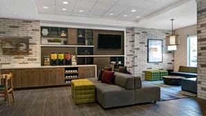 A modern living room with a built-in kitchen, a large flat-screen TV, and a cozy seating area with a green ottoman and a grey sectional sofa.