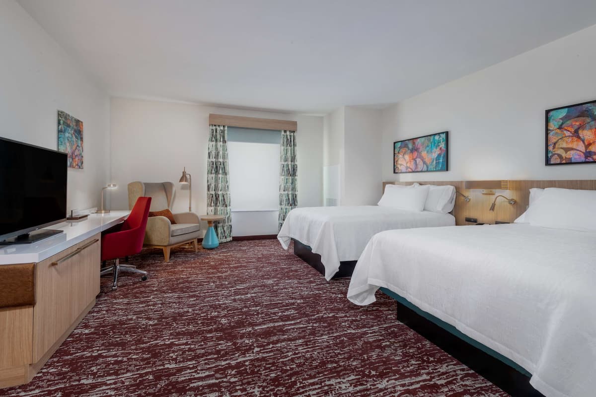 A hotel room with a desk, two beds, a TV, and artwork on the walls.