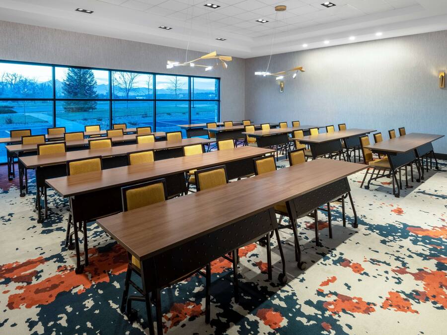 Un salón de conferencias con varias mesas y sillas, amplias ventanas que dan a vistas de árboles y montañas, y una alfombra estampada.