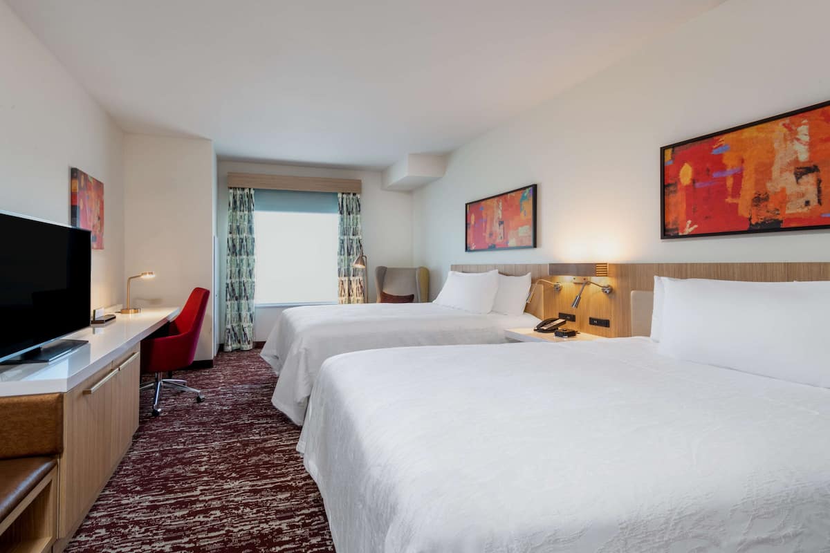 A hotel room with two beds, a desk with a TV, a red chair, and artwork on the walls.
