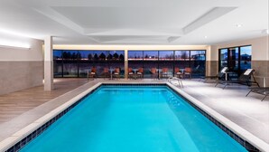 A modern indoor swimming pool with clear blue water, surrounded by wooden flooring and furnished with tables and chairs.