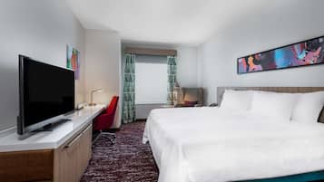 A hotel room with a bed, a TV, a desk with a chair, a window with curtains, and a painting on the wall.