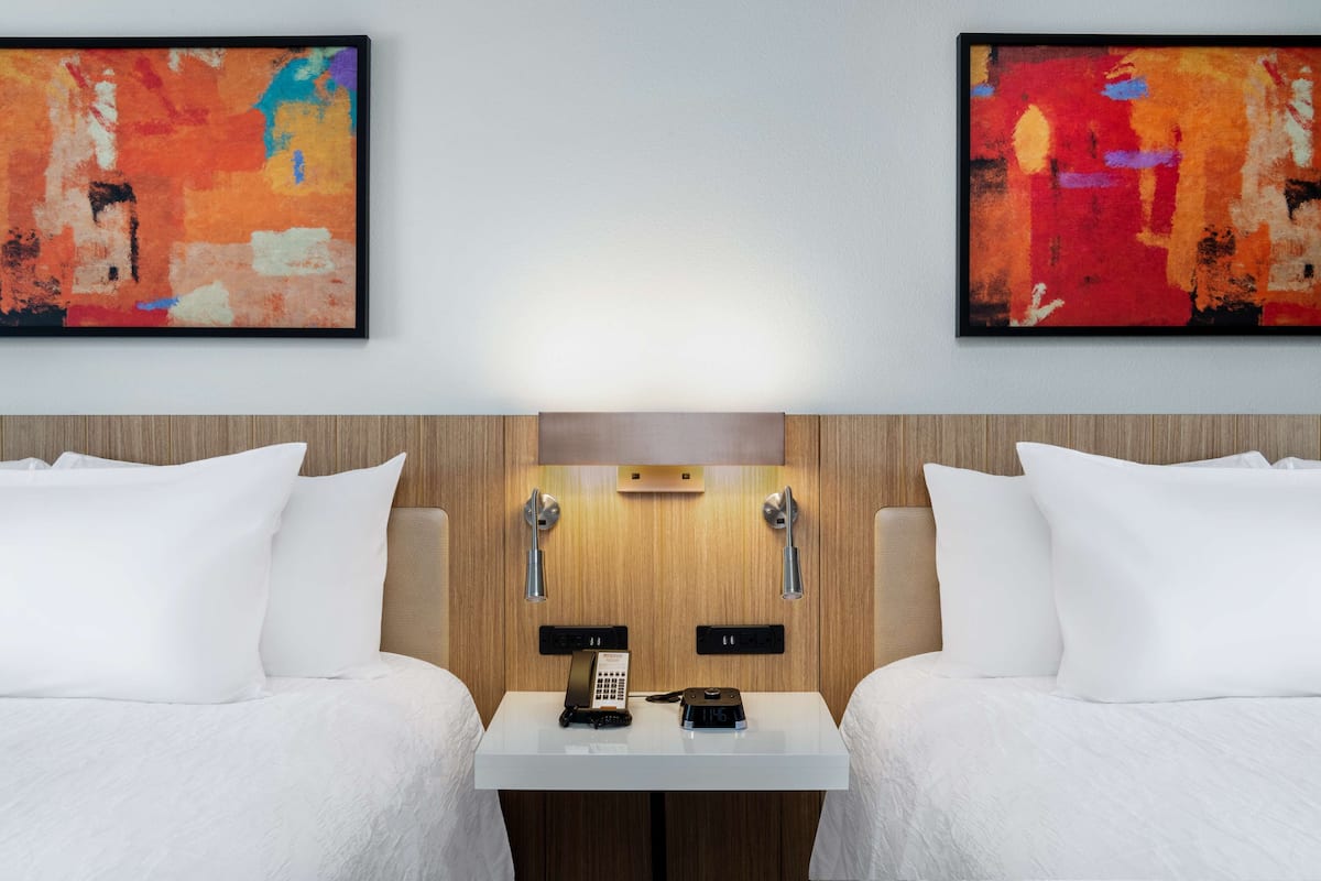 A hotel room with two beds, a wooden headboard, a bedside table with a phone and a lamp, and two abstract paintings on the wall.