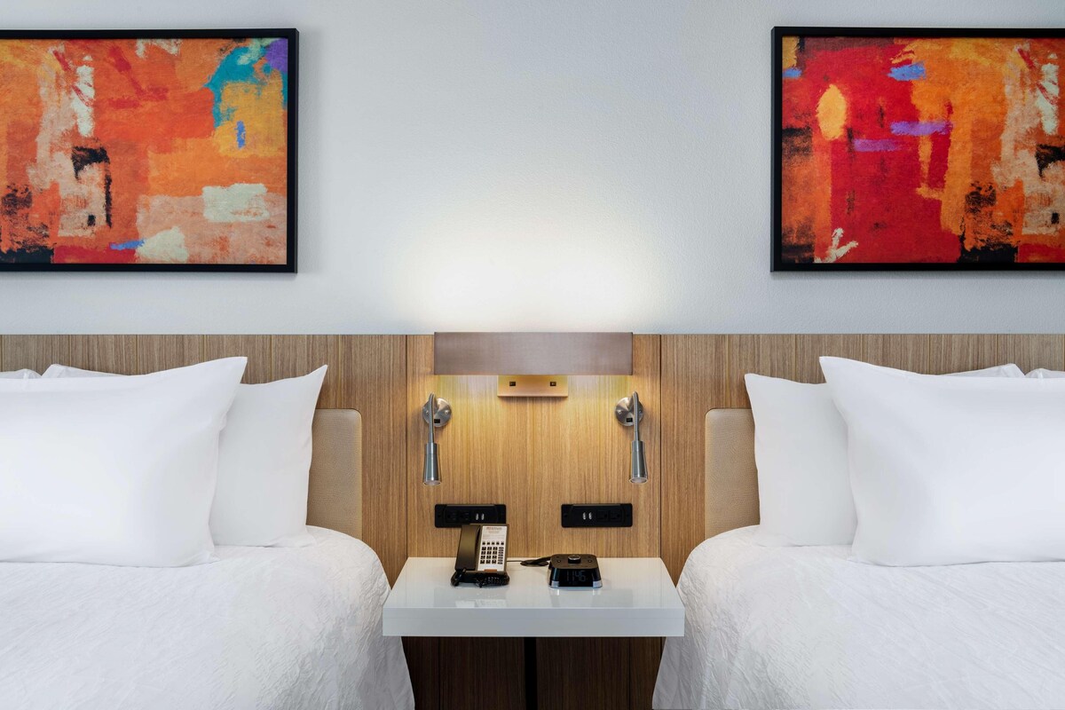A hotel room with two beds, a wooden headboard, a small table with a phone and a remote, and two abstract paintings on the wall.