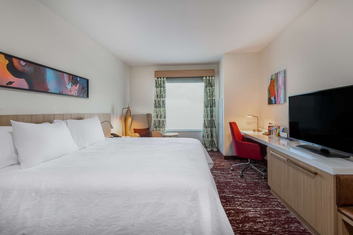 A hotel room with a large bed, a desk with a computer, a TV, and a window with curtains.