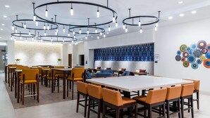 A modern dining area with a large rectangular table surrounded by orange chairs, overhead circular lighting, and a decorative wall with a blue pattern.