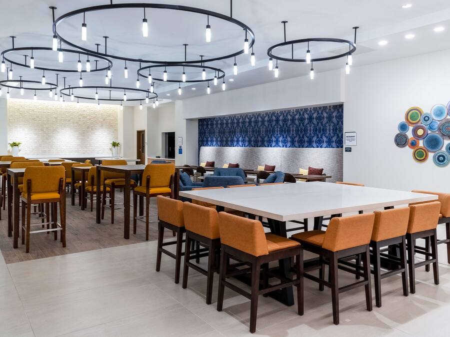 A modern dining area with a large rectangular table surrounded by orange chairs, overhead circular lighting, and a decorative wall with a blue pattern.