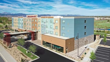A multi-story hotel with a modern design, featuring a mix of light and dark facades, a parking lot, and a view of mountains in the background.