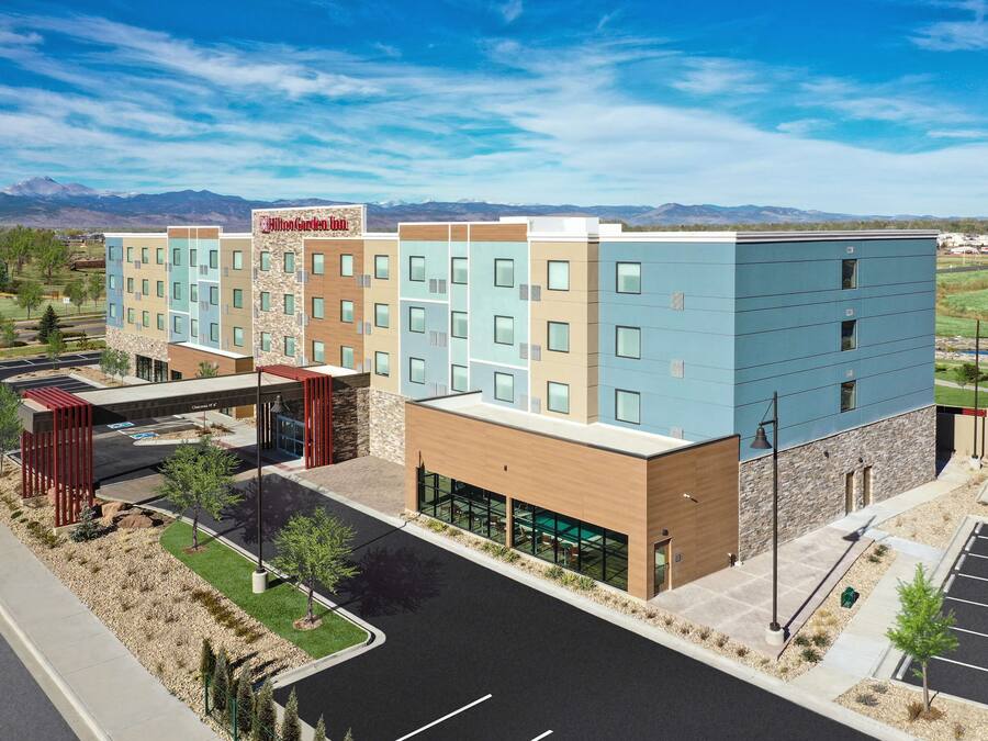 A multi-story hotel with a modern design, featuring a mix of light and dark facades, a parking lot, and a view of mountains in the background.