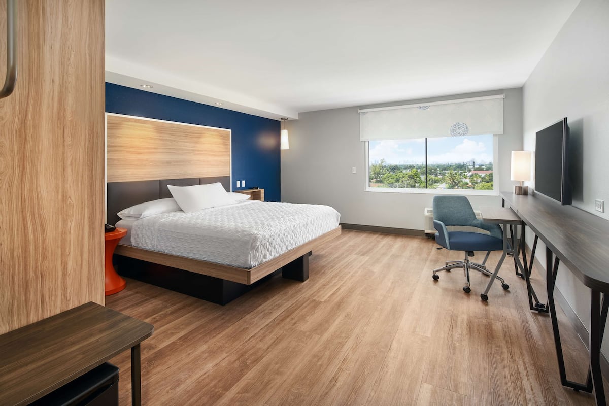 A modern hotel room with a bed, desk, chair, and a large window with a city view.