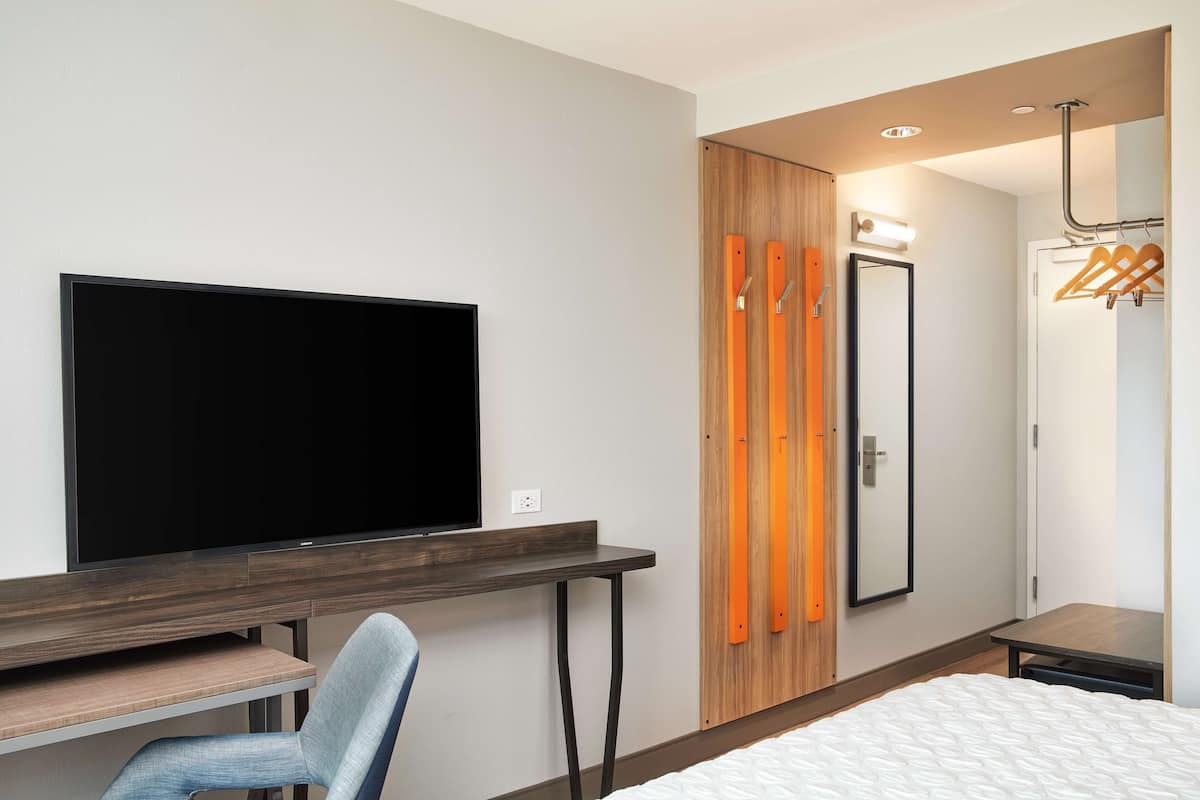 A modern hotel room with a flat-screen TV, a desk, a chair, a bed, a wardrobe with a mirror, and a coat rack.